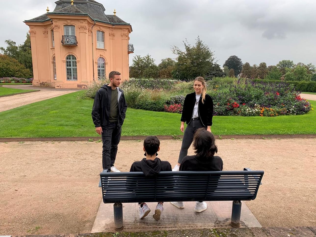 Two young people on a park bench, two staff members of the youth work are standing in front of it, in the background you can see the Pagoda Castle