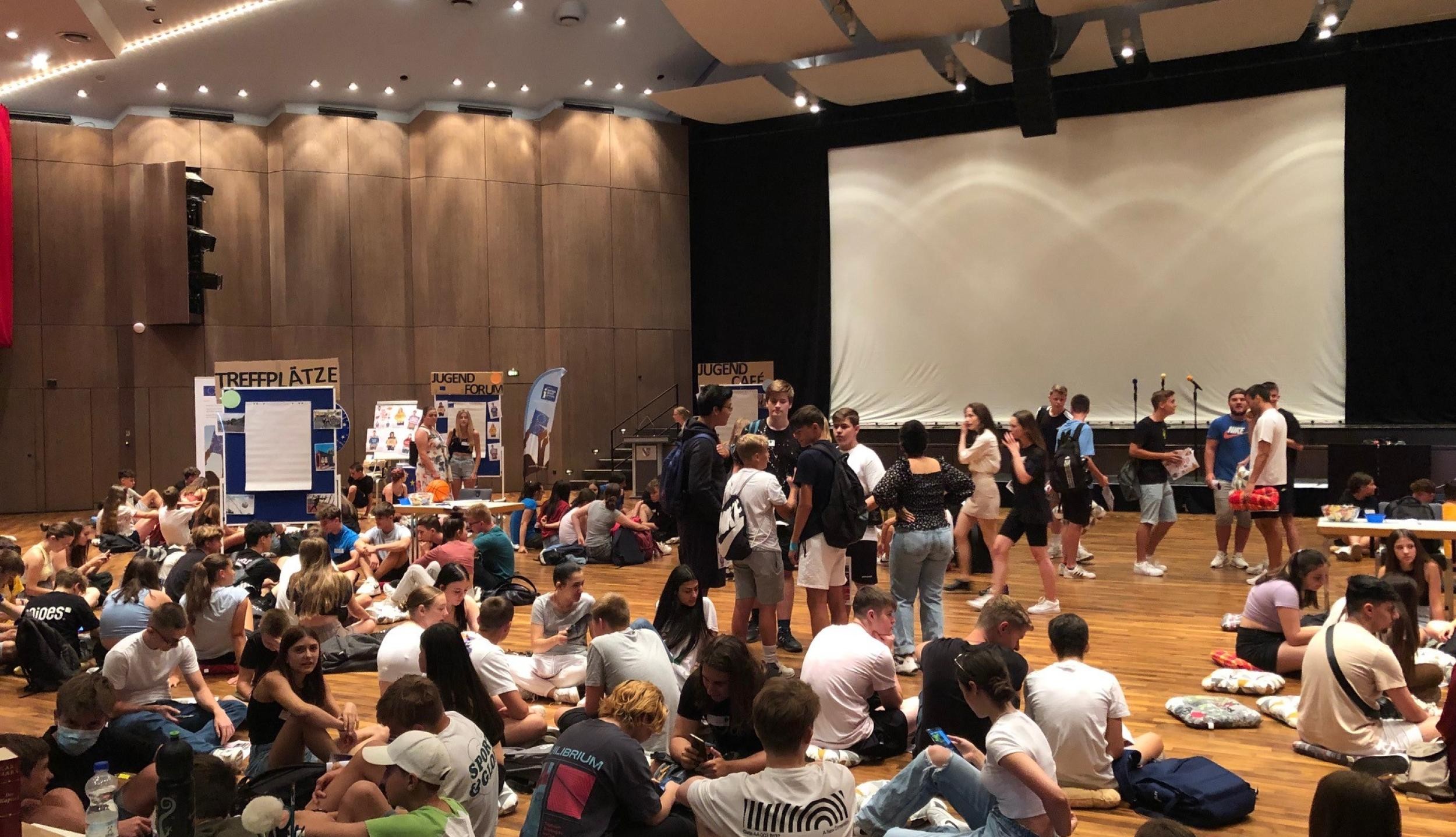 Many young people sit and stand in a large hall. There are information stands and a large screen in the background.