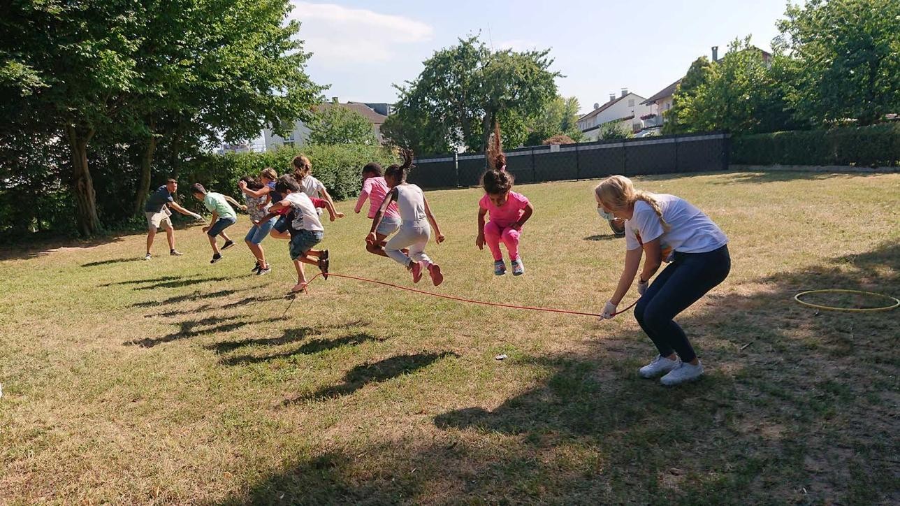 Several children jumping over a skipping rope on a meadow, the rope is swung by two adults, in the background you can see houses and trees
