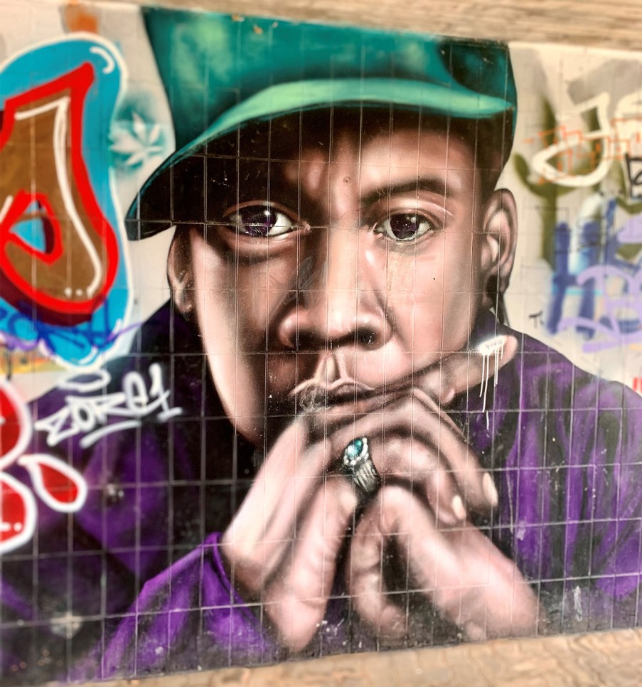 Mur recouvert de graffitis colorés représentant la tête, les épaules et les mains d'un homme portant un bonnet vert, une veste violette et une bague au doigt. La tête est appuyée sur les mains jointes.