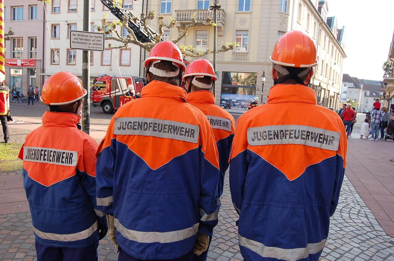 Jugendfeuerwehr Rastatt Maibaum. Foto: Freiwillige Feuerwehr Rastatt Vier Jugendliche mit Feuerwehruniform von hinten, im Hintergrund Häuser, Straßen, Zuschauer, ein Drehleiterfahrzeug der Feuerwehr