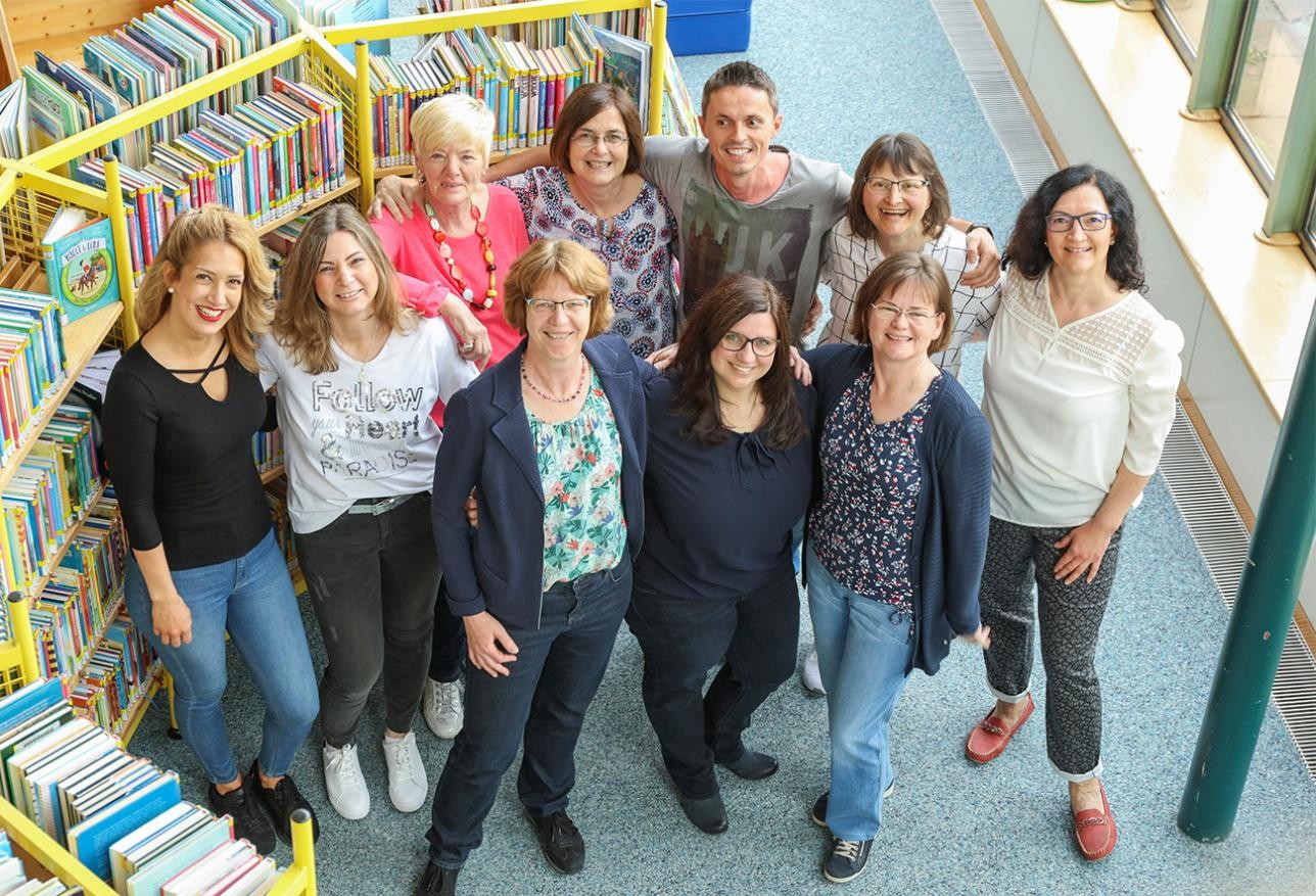Group photo of the city library staff, 10 women and 1 man, in the background shelves with books. 