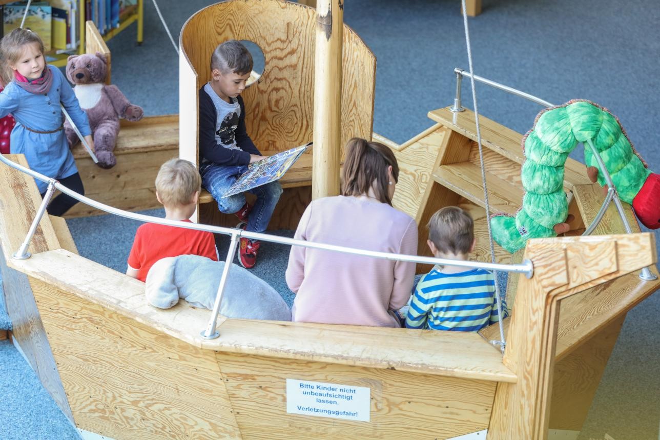 Several children and a mother sit on a wooden bench modeled on a boat and read books.
