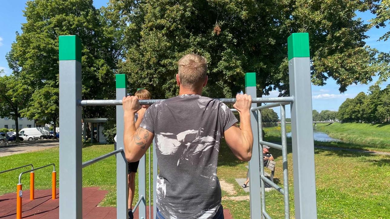 A man from behind does a pull-up on a bar of the calisthenics facility, in the background trees and meadow