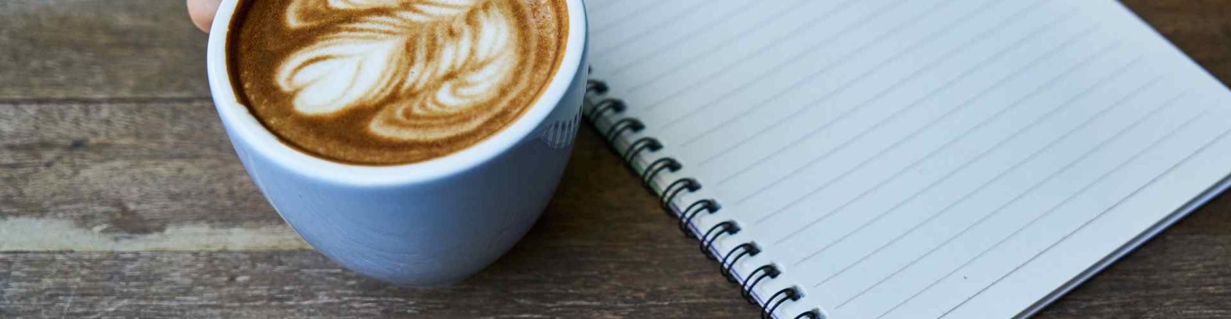 Eine Tasse mit Kaffee und ein Schreibblock mit Linien auf einem Holztisch, eine Hand hält die Tasse am Henkel fest