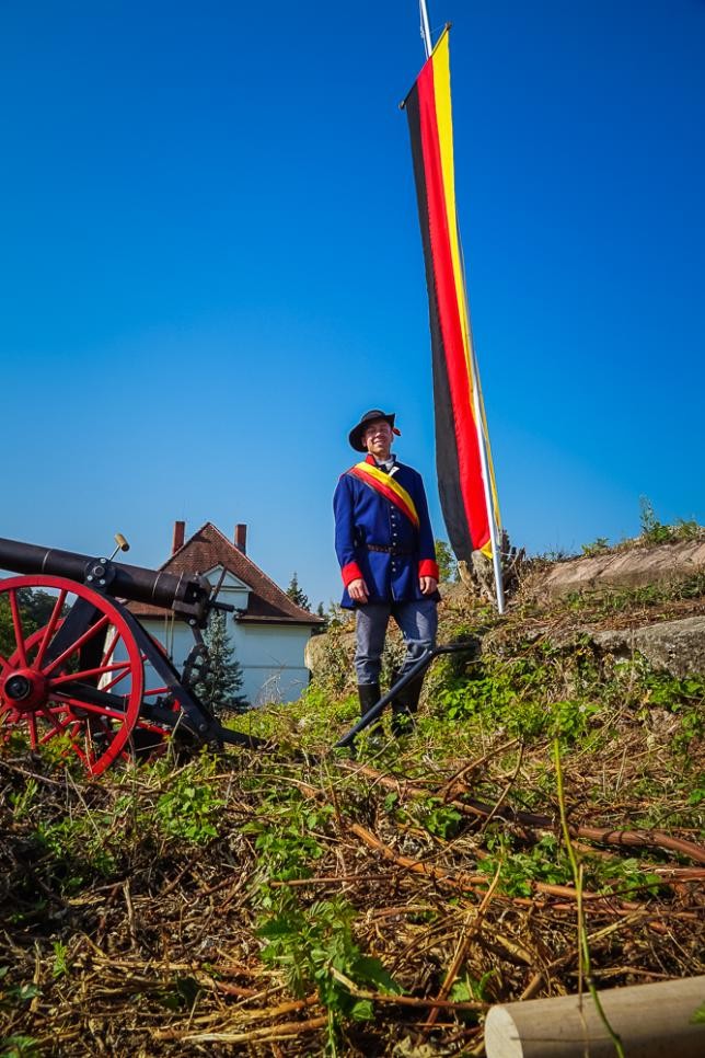Schauspieler in historischem Kostüm steht vor einer Fahne und einer Kanone