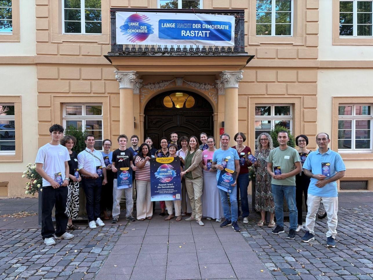 Am 2. Oktober feiert Rastatt die „Lange Nacht der Demokratie“. Das vielfältige Programm wird überwiegend von ehrenamtlich Engagierten zusammengestellt und organisiert. Foto: Stadt Rastatt/Heike Bornhäußer Menschen vor dem Rathaus mit Plakat "Lange Nacht der Demokratie"