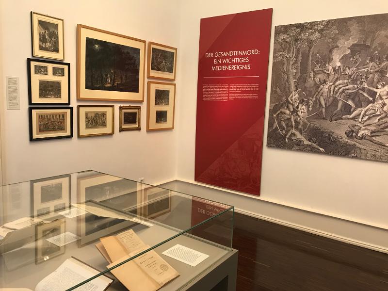 Sichtbar ist ein Teil der Vitrine mit Schriftstücken und verschiedene Bilder an den Wänden, welche den Gesandtenmord dokumentieren
