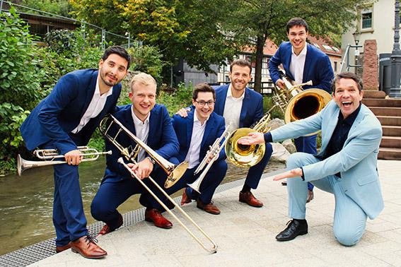 5 Männer in blauem Anzug und Blechblasinstrument, im Vordergrund ein Mann im hellblauen Anzug, der auf die anderen zeigt.