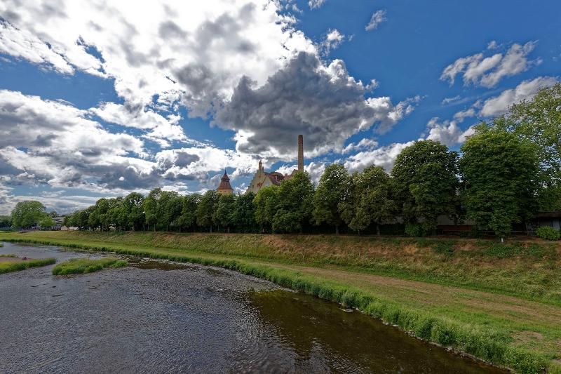 im Vordergrund die Murg, dahinter Bäume und Wasserturm und Brauereigebäude, Wolken