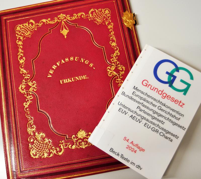 Links ist die 25-jährige Jubiläumsausgabe der badischen Verfassung aus dem Jahr 1818 zu sehen. Rechts ist das deutsche Grundgesetzbuch aus dem Jahr 2024.