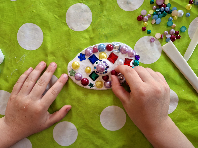Kinderhände verzieren eine Tonscheibe mit bunten Perlen und Glassteinen