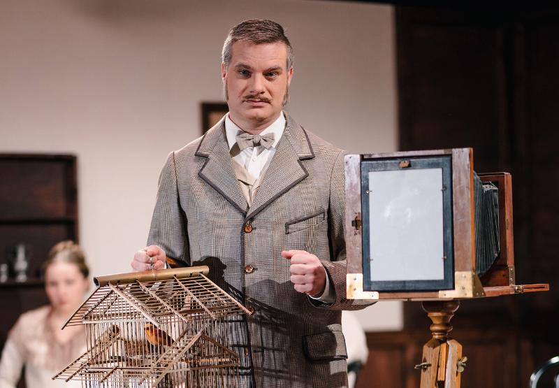 Herr in grauem Anzug mit Vogelbauer in der Hand. Im Hintergrund eine Lochkamera.
